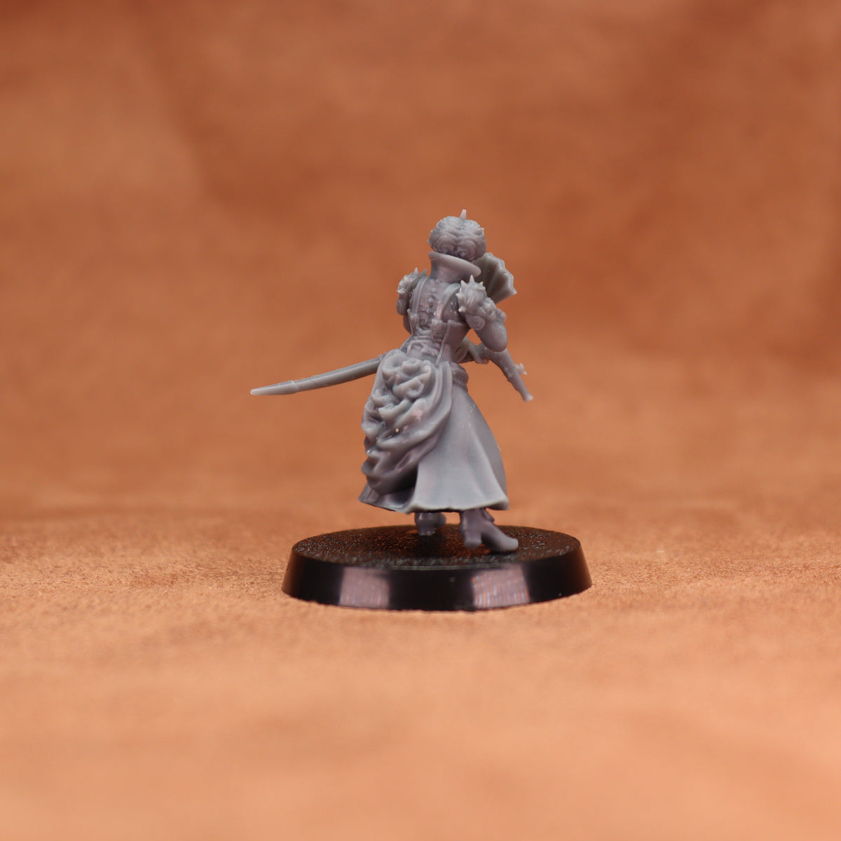 A small unpainted gray fantasy miniature figurine of a female warrior holding a sword, standing on a round black base against a warm brown textured background.