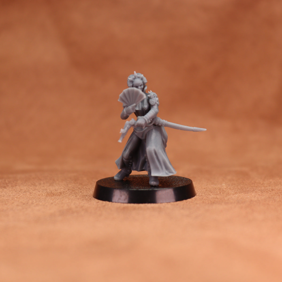 A small unpainted gray fantasy miniature figurine of a woman holding a sword and folding fan, standing on a round black base against a warm brown textured background.