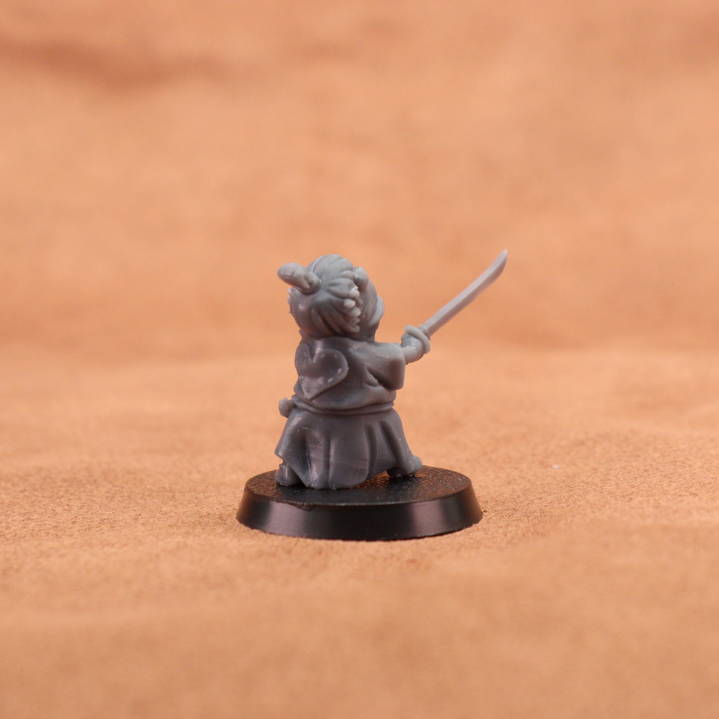 A small gray fantasy miniature figurine of a child warrior holding a sword, standing on a round black base against a warm brown textured background.