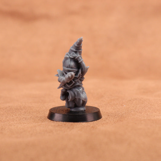 A small unpainted gray fantasy miniature figurine of a wizard or gnome-like character standing on a round black base, photographed against a warm tan textured background.