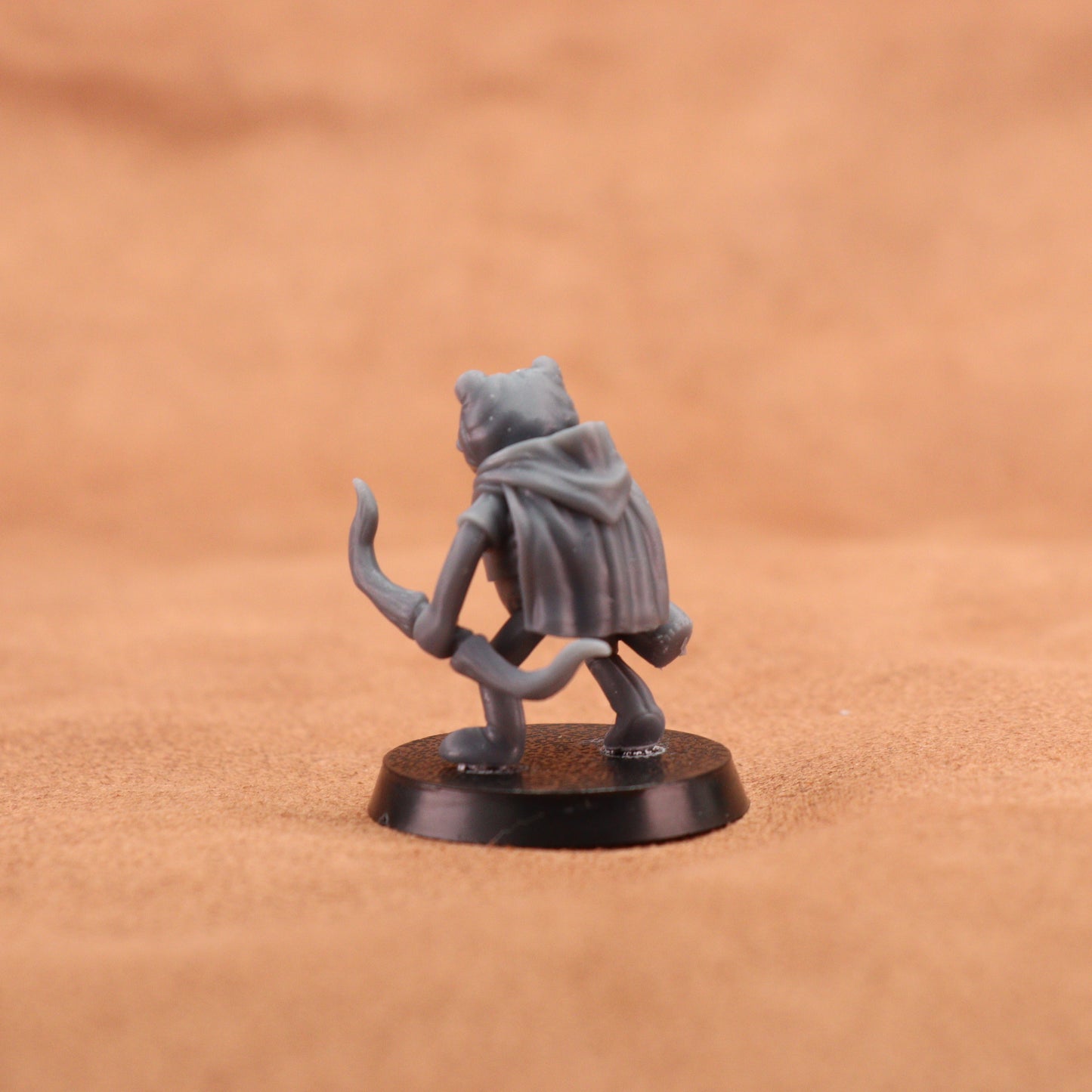 A small gray fantasy miniature figurine of a cat-like character wearing a hooded cloak, standing on a round black base against a warm tan textured background.