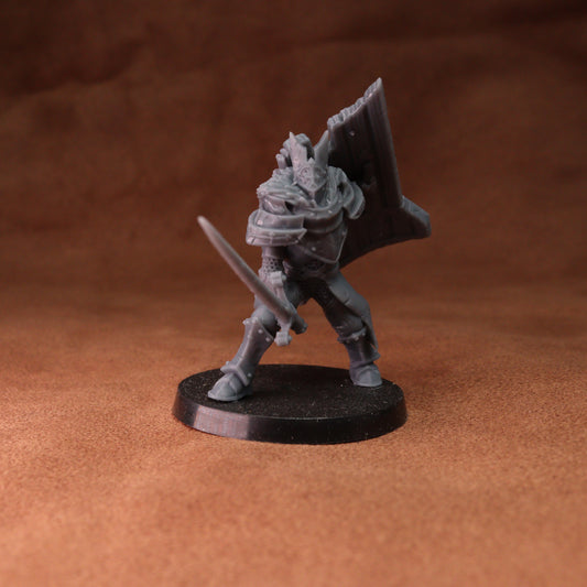 An unpainted gray fantasy warrior miniature figurine holding a sword and shield, standing on a round black base against a textured brown background.