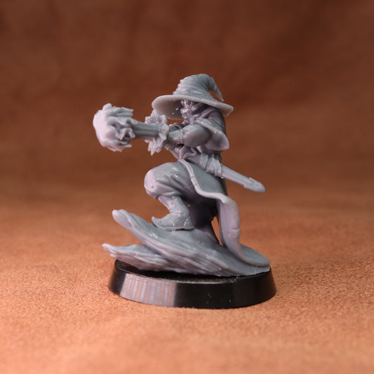 A gray unpainted fantasy miniature figurine of a wizard casting a spell, mounted on a round black base, set against a warm brown textured background.