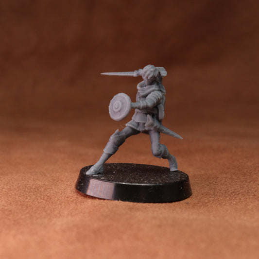 An unpainted gray fantasy warrior miniature figurine holding a sword and shield, standing on a round black base against a textured brown background.
