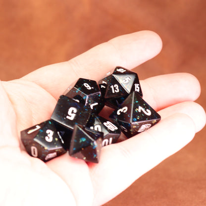 Hand holding a set of black polyhedral dice with white numbers against a brown background