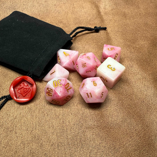 Pink and white dice set with gold numbers on a beige surface, accompanied by a black drawstring bag.