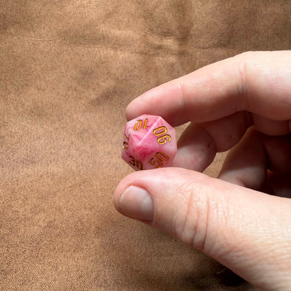 Hand holding a pink die with gold numbers on a brown background