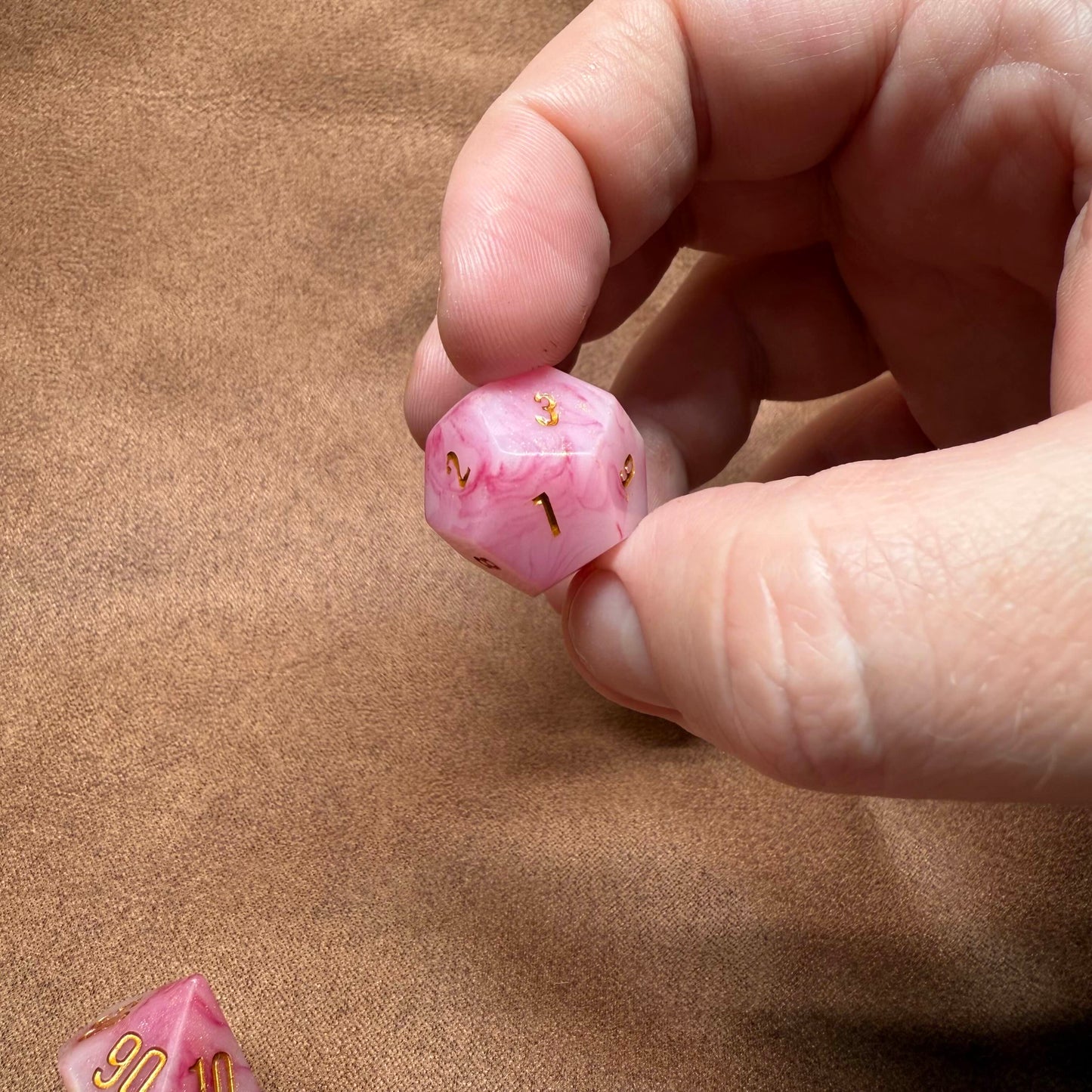 Hand holding a pink die with gold numbers on a brown background
