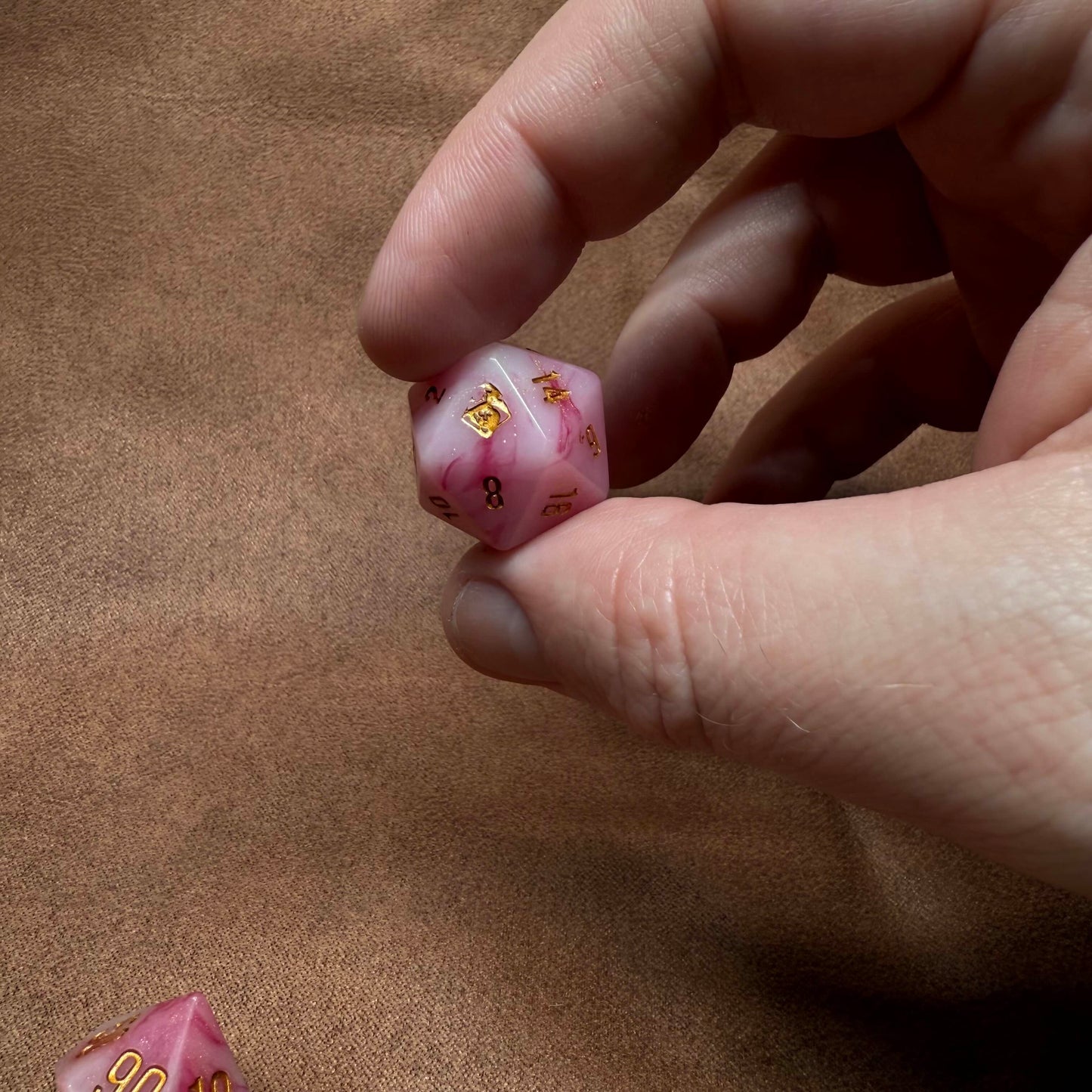 Hand holding a pink die with gold text on a brown surface