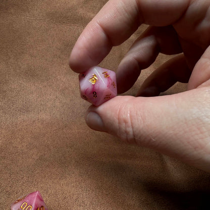 Hand holding a pink die with gold text on a brown surface