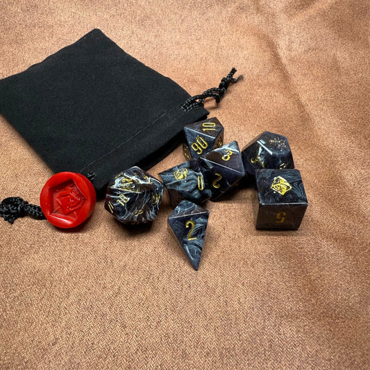 Set of black dice with gold numbers on a brown surface next to a black drawstring bag and red die.