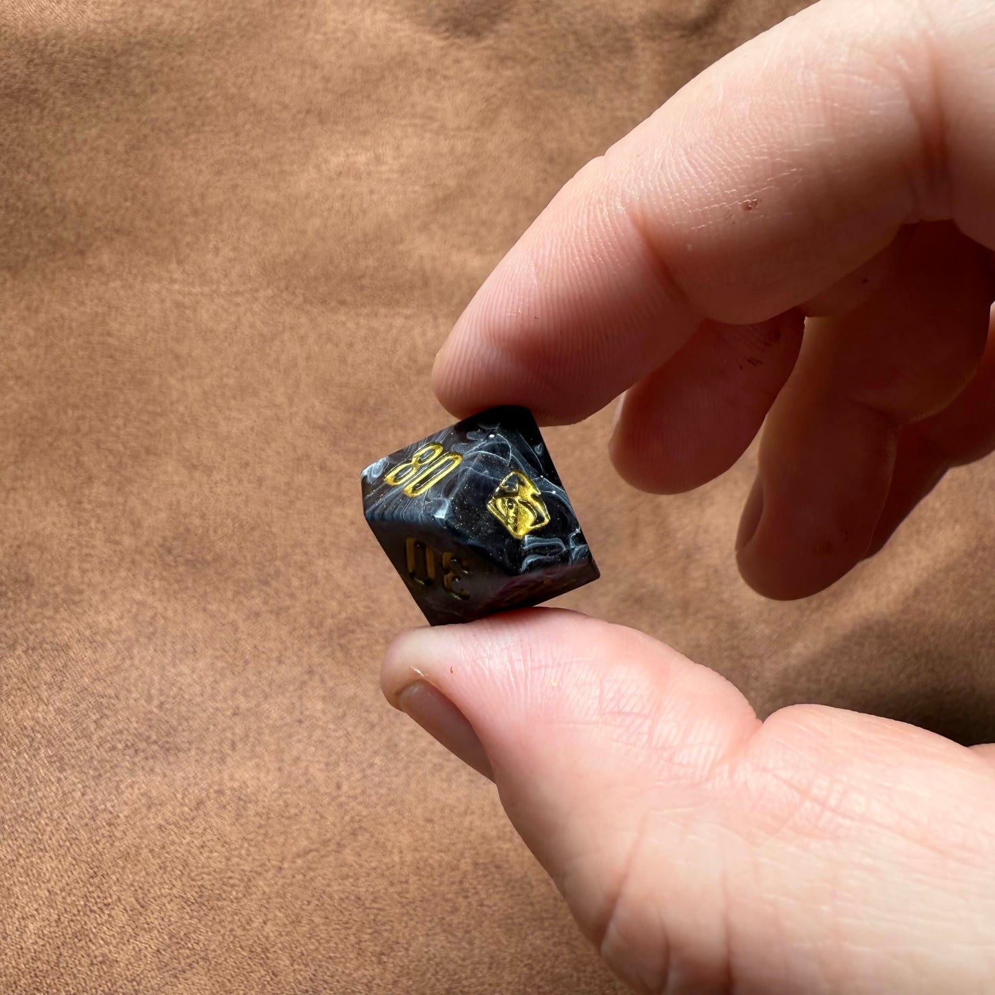 Hand holding a black dice with gold numbers on a brown leather surface