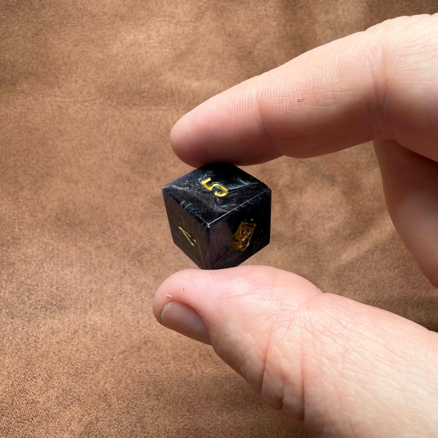 Hand holding a small black die with gold numbers on a brown background