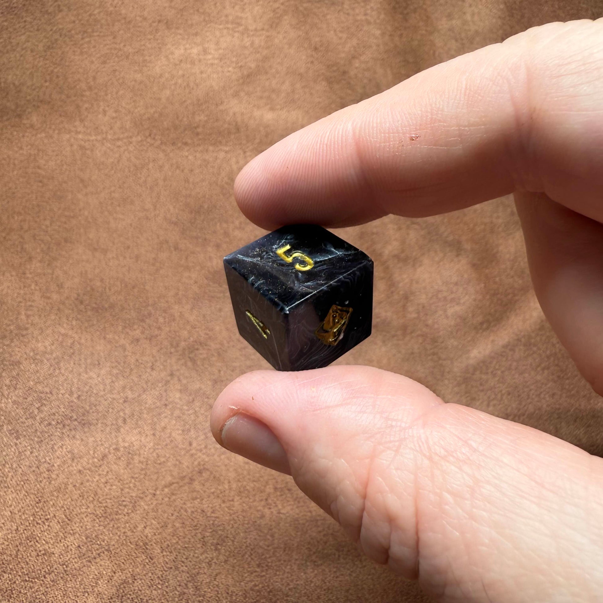 Hand holding a small black die with gold numbers on a brown background