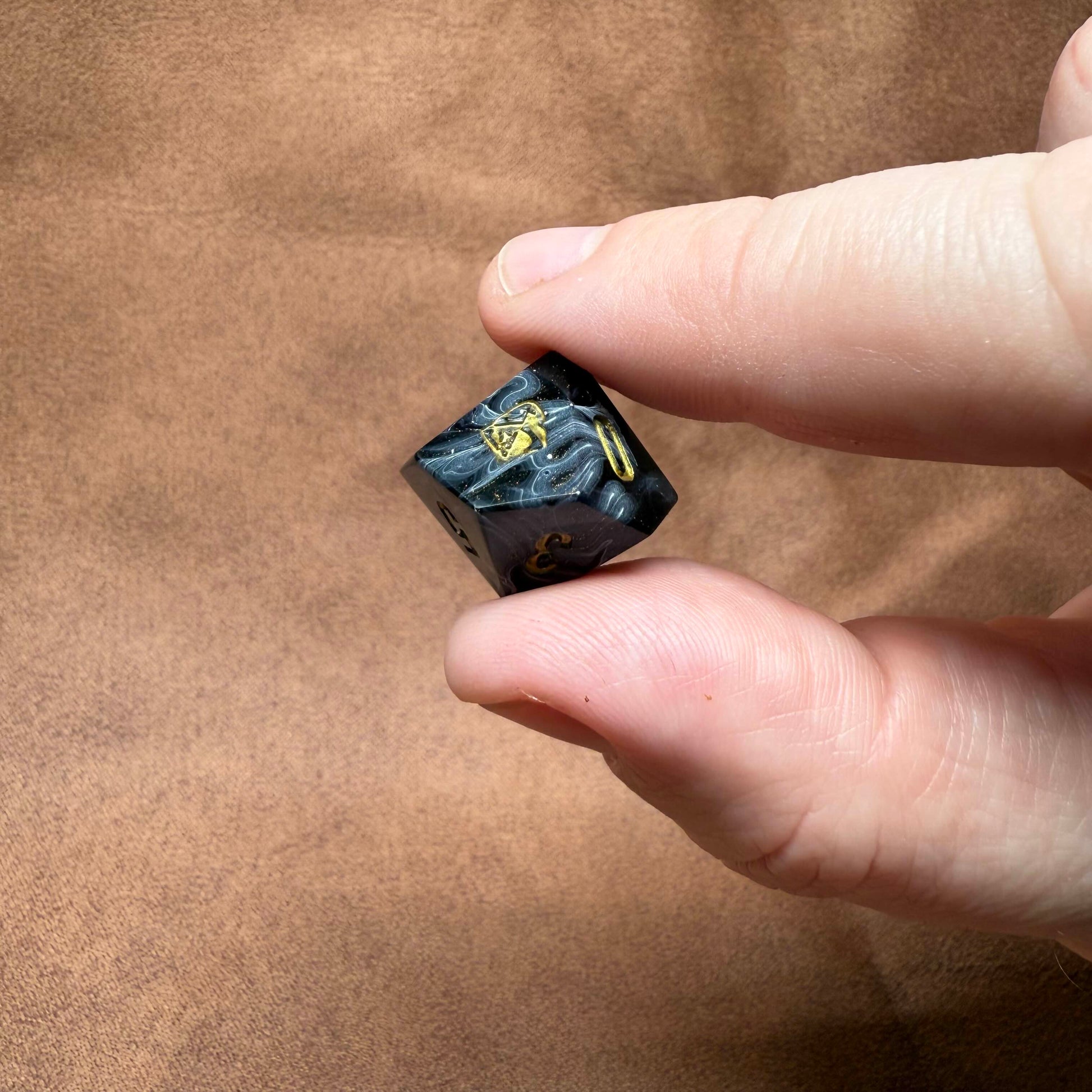 Hand holding a small black die with yellow text on a brown background