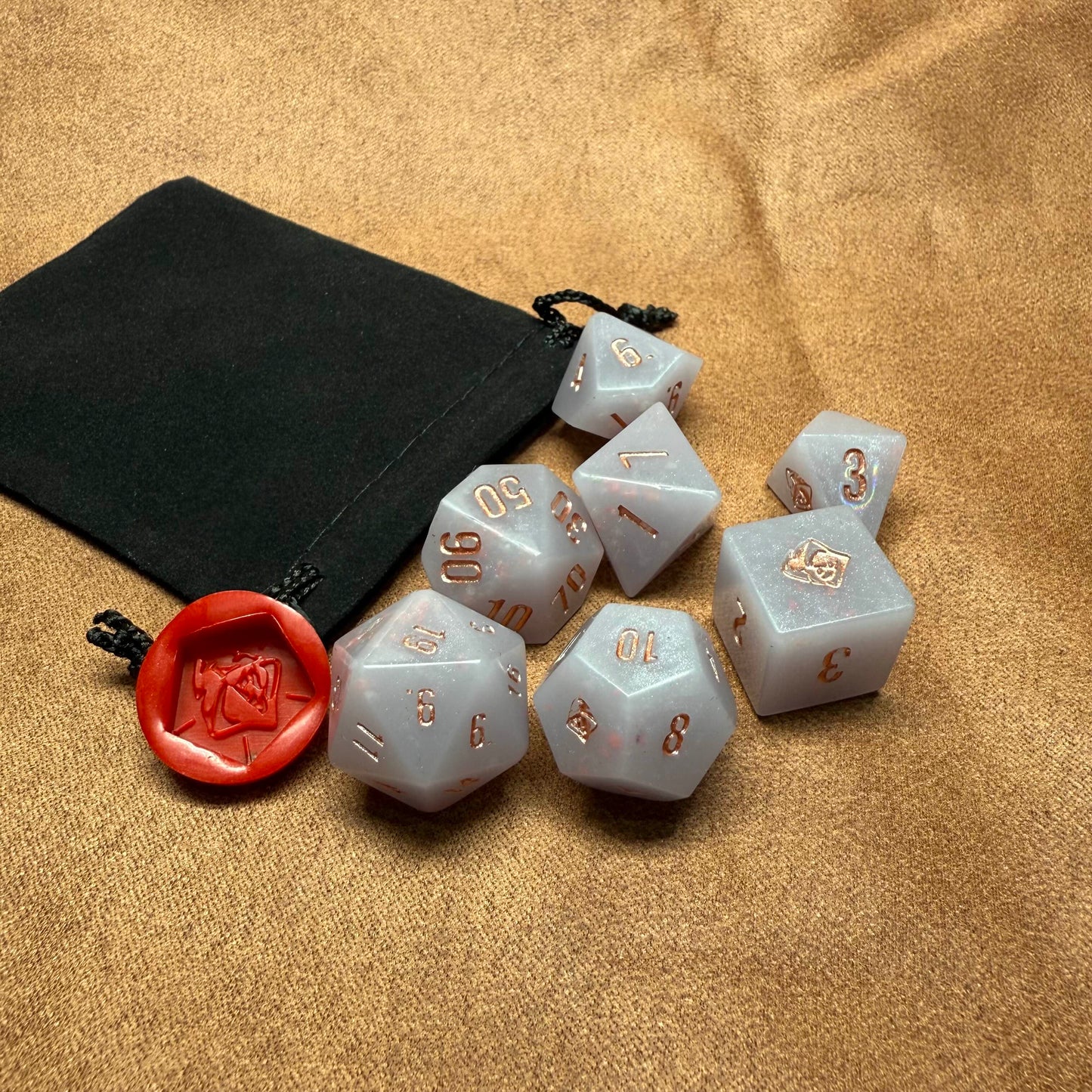 Set of polyhedral dice with gold numbering on a beige surface with a black drawstring bag.