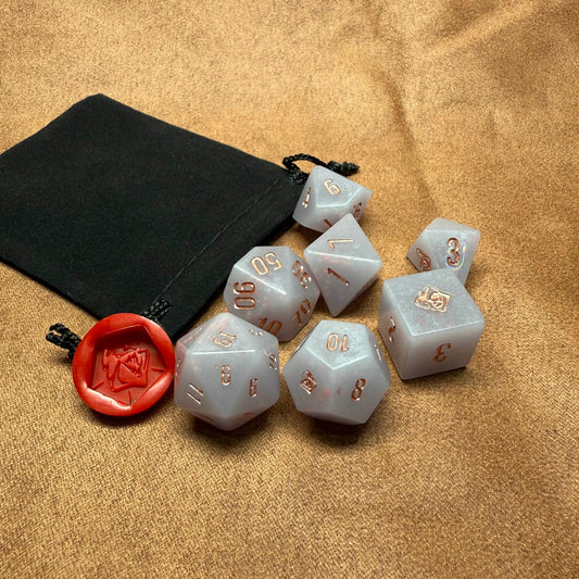 Set of polyhedral dice with gold numbering on a beige surface with a black drawstring bag.