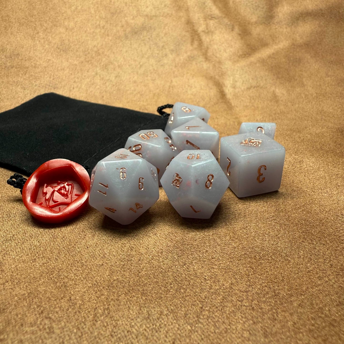 Set of light blue polyhedral dice with gold numbers on a beige surface, accompanied by a red die and black pouch.