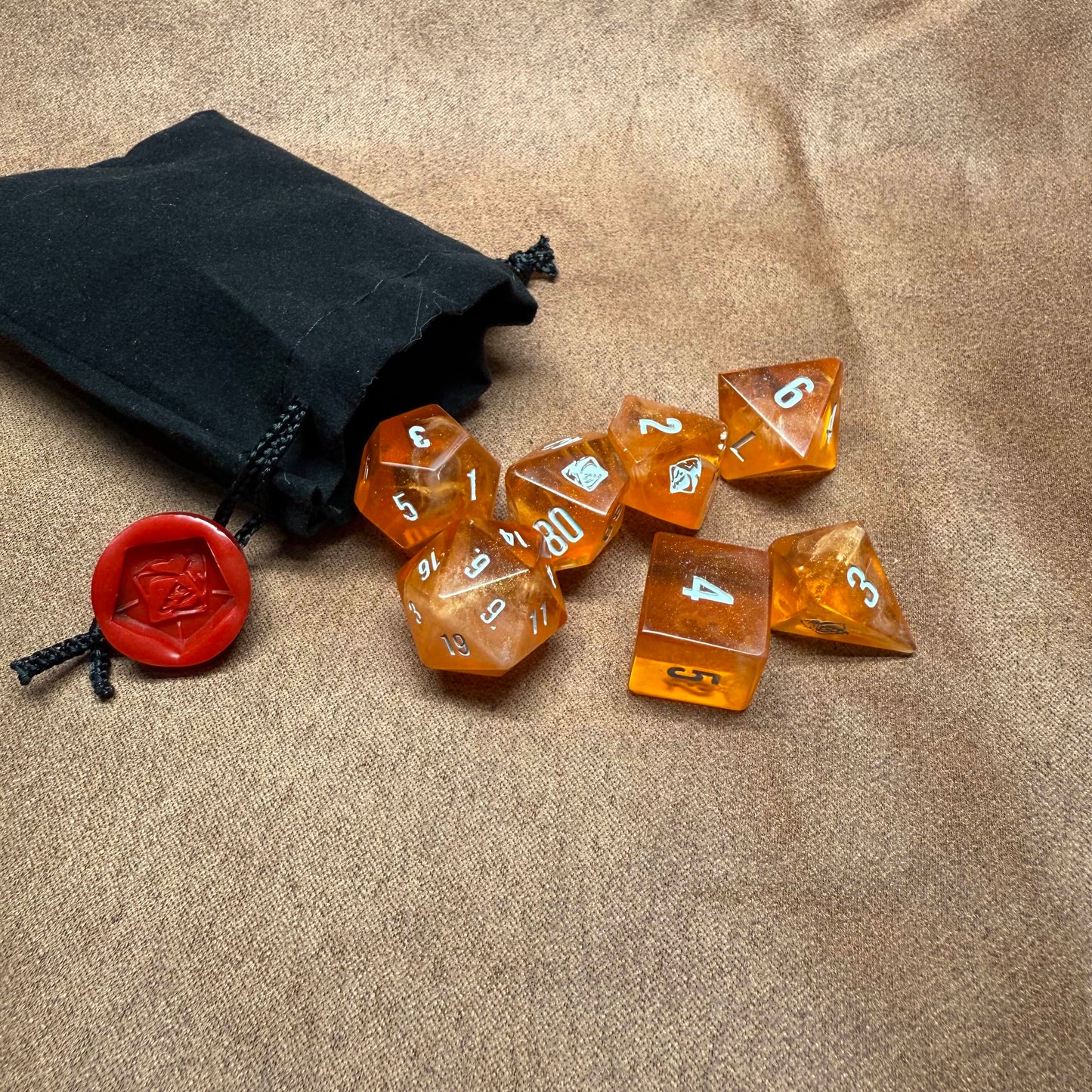Set of amber-colored polyhedral dice with numbers on a beige surface next to a black drawstring bag and red die with a seal.