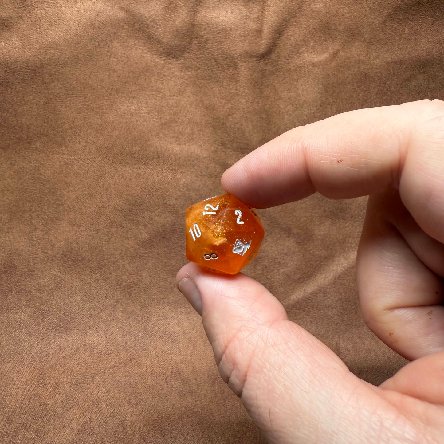 Hand holding a small orange die with numbers on a brown textured background