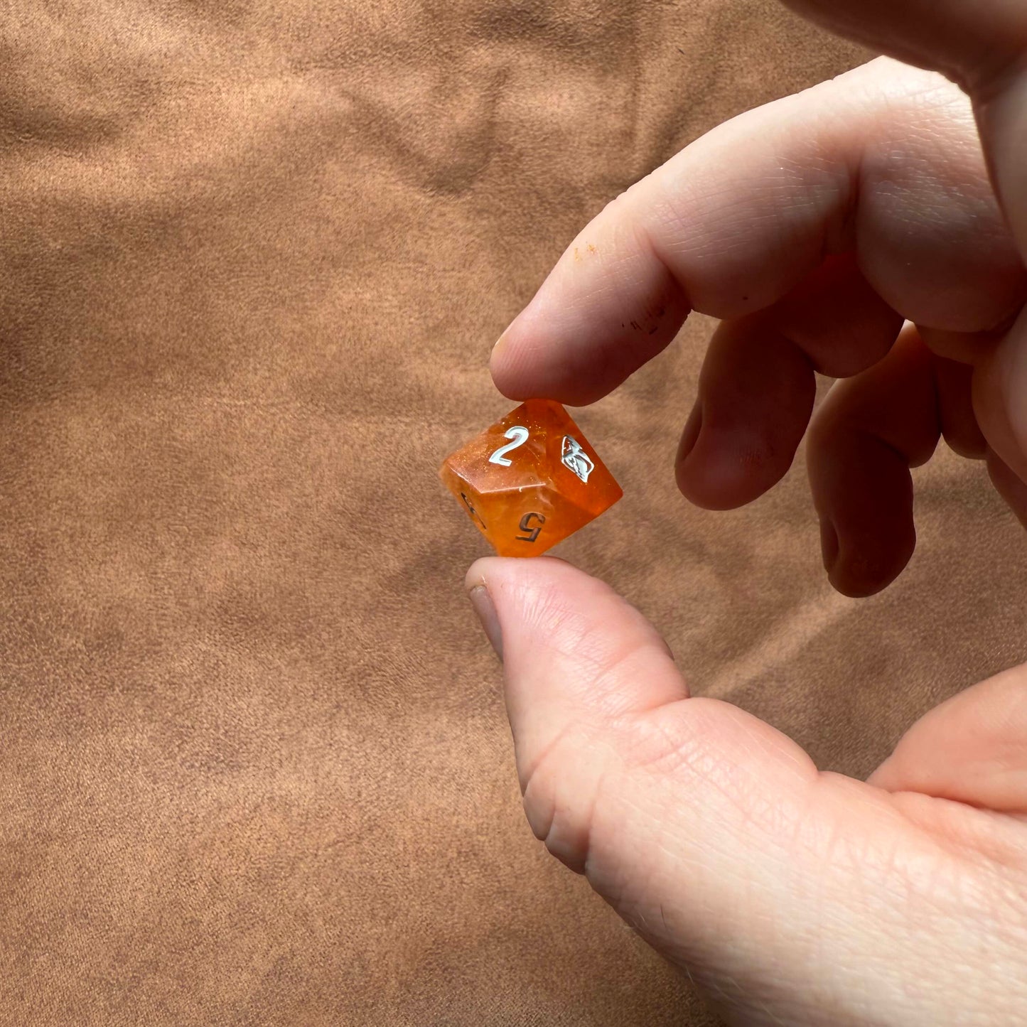 Hand holding a small orange die with white numbers on a brown textured surface