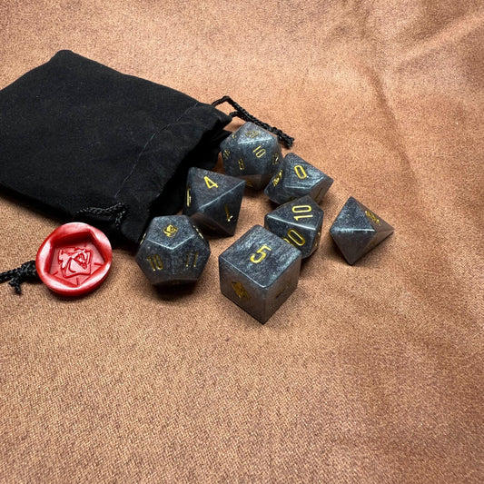 Set of gray polyhedral dice with gold numbers on a brown surface, accompanied by a black drawstring bag and a red die.