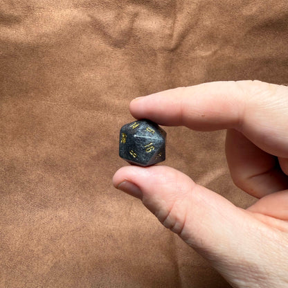 Hand holding a black polyhedral dice with yellow numbers on a brown leather surface