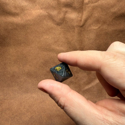 Hand holding a small black and gold die against a brown fabric background