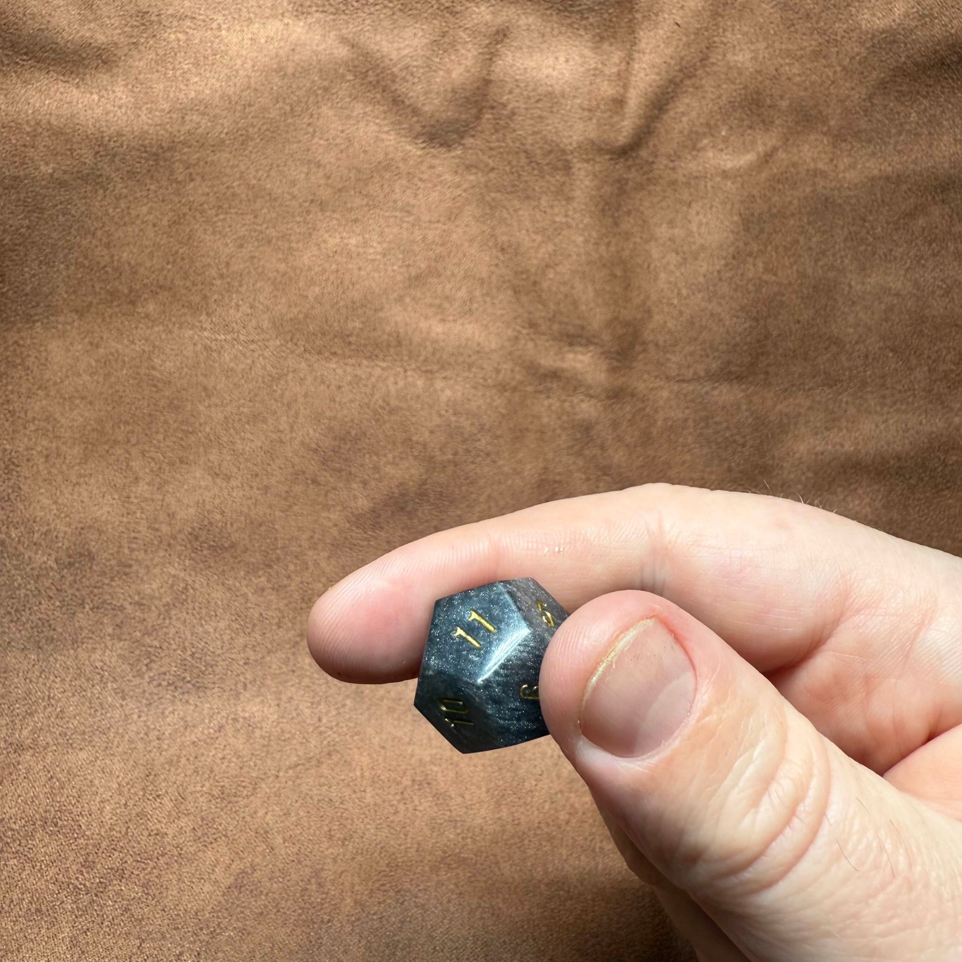 Hand holding a small black die with gold numbers on a brown fabric background