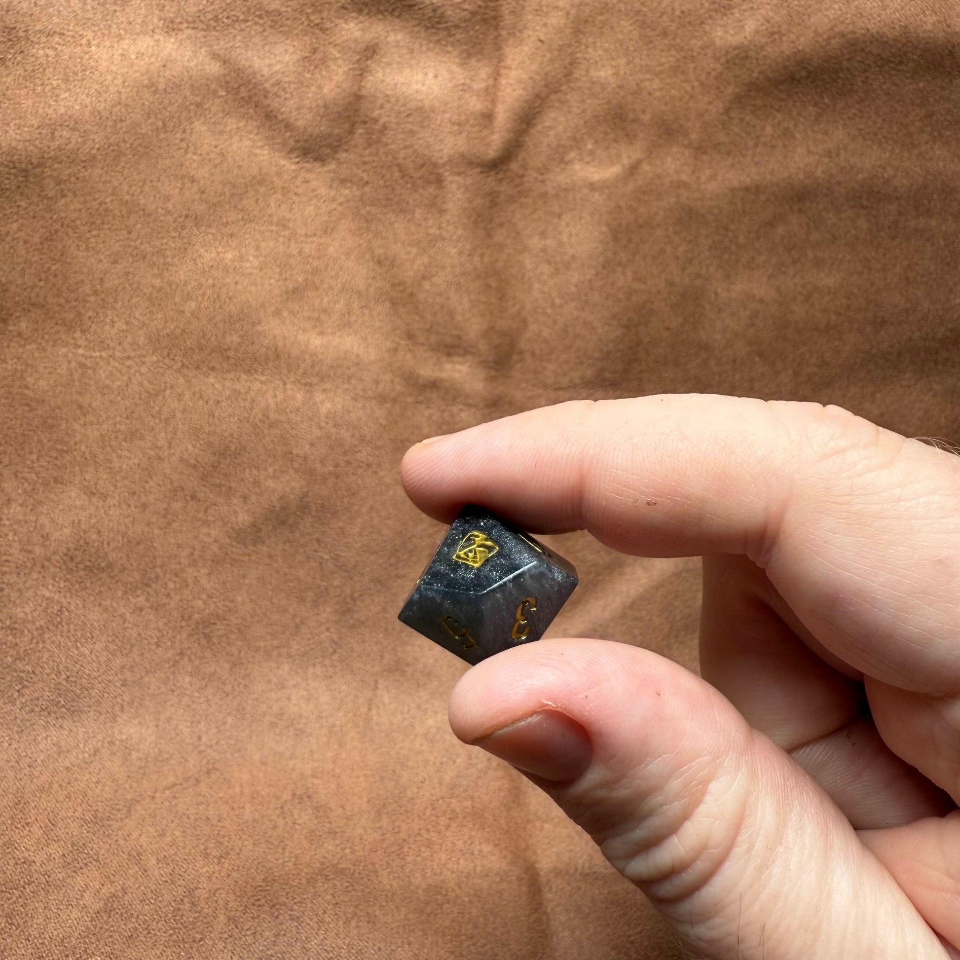 Hand holding a small black die with gold symbols against a brown leather background