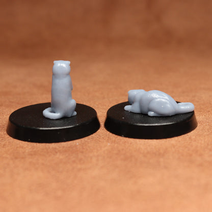 Two small figurines of a standing and a lying cat on black bases against a brown background