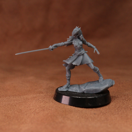 A gray unpainted fantasy miniature figurine of a female warrior holding a sword, standing on a black circular base against a brown textured background.