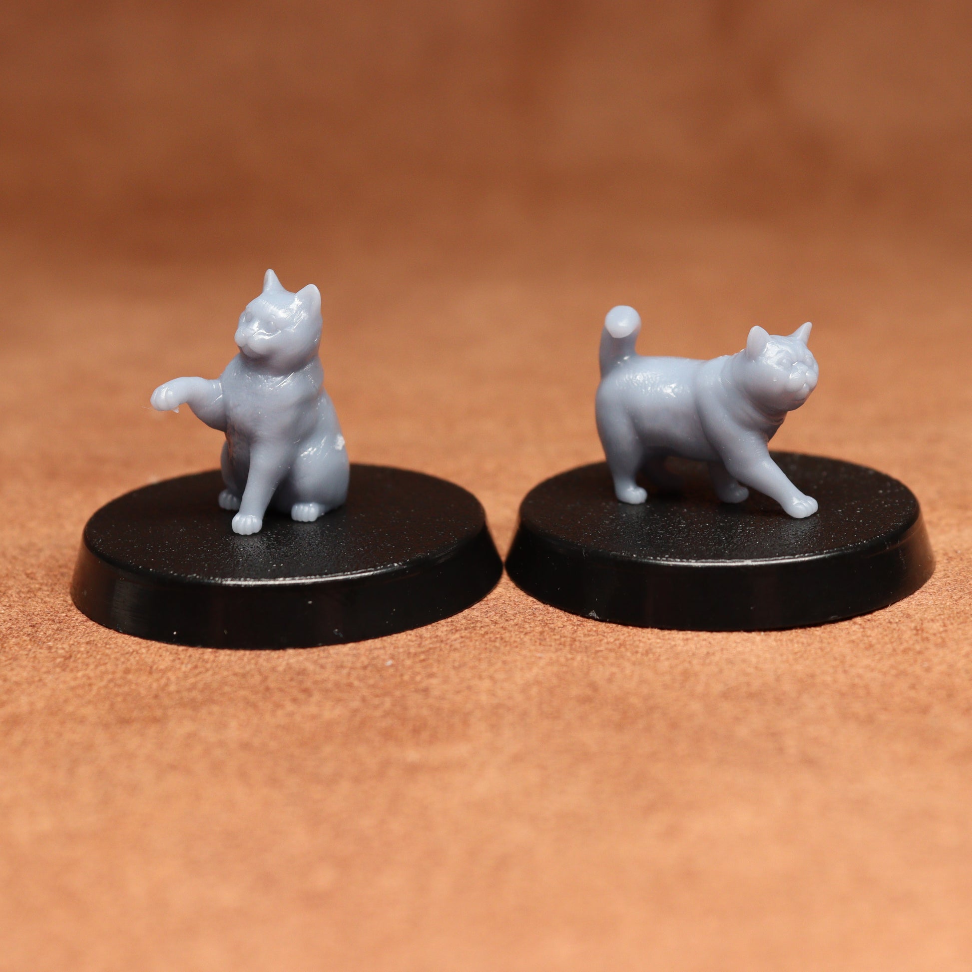 Two miniature cat figurines on black round bases against a brown background