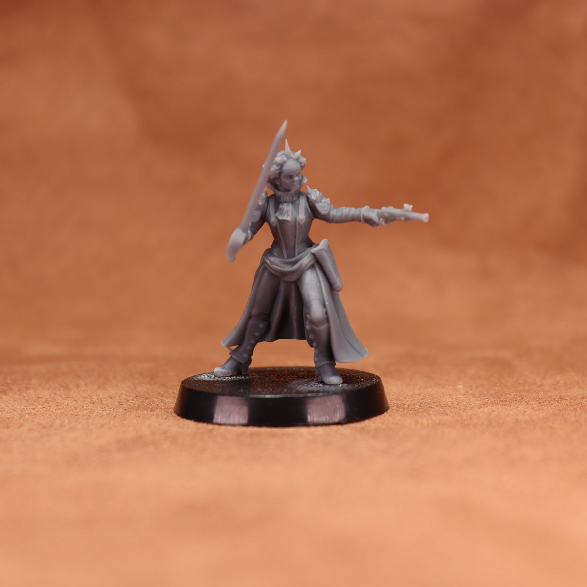 A gray unpainted fantasy tabletop miniature figurine of a female warrior holding a sword and pistol, standing on a round black base against a warm brown textured background.