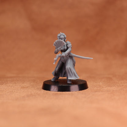 A small unpainted gray fantasy miniature figurine of a woman holding a sword and folding fan, standing on a round black base against a warm brown textured background.