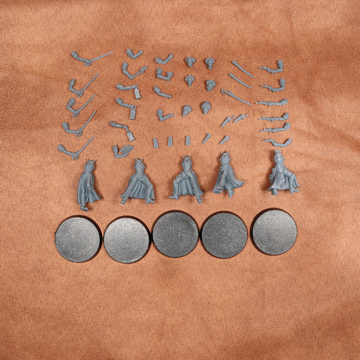 A set of unpainted miniature figurines and accessories for tabletop gaming, including four small character figures with detailed robes and multiple weapon and accessory pieces, arranged on a textured tan leather background. Five round gray bases are shown at the bottom.