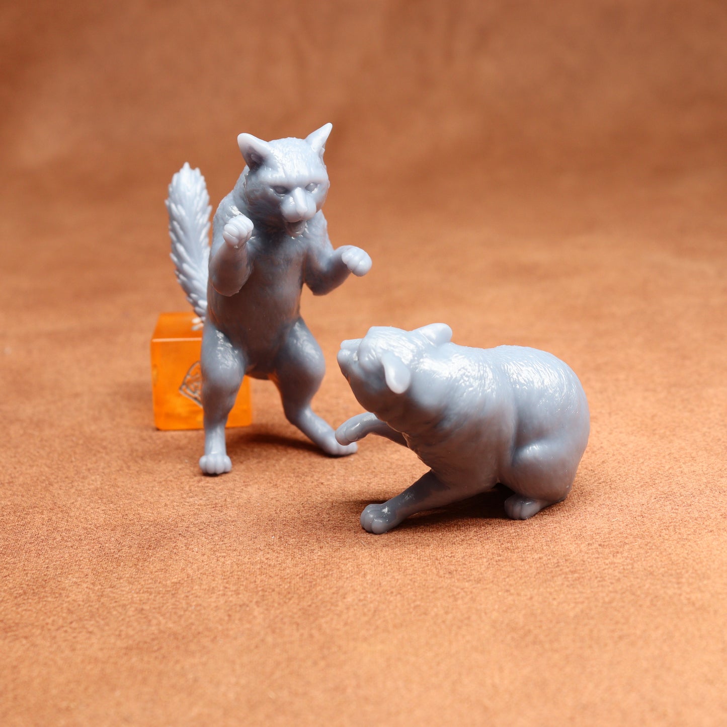 Two gray cat figurines on a brown surface with an orange dice in the background.