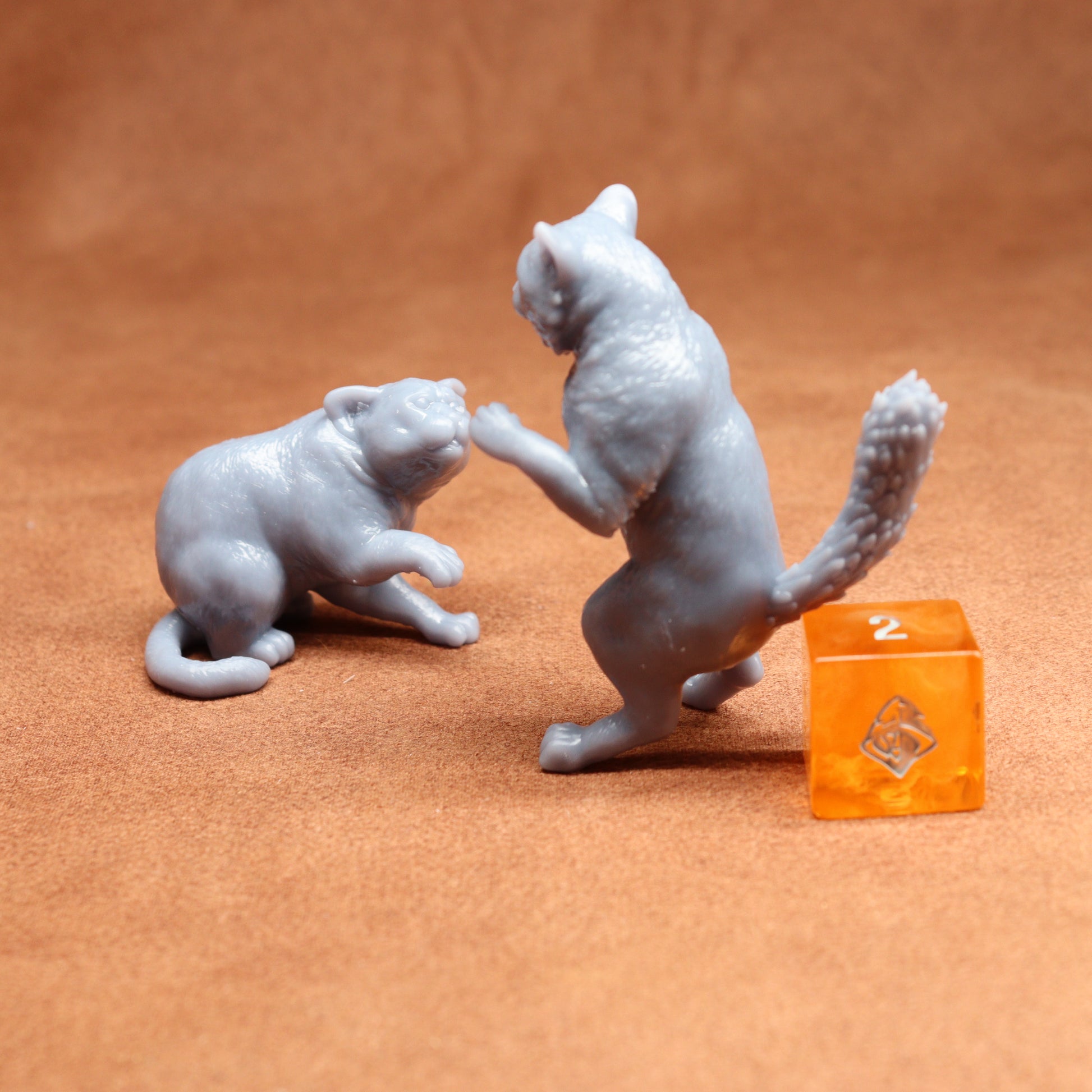 Two grey cat figurines on a brown surface with an orange dice in the background.