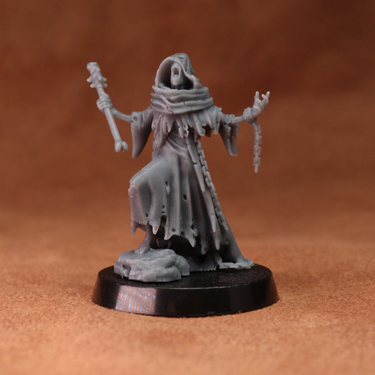 A small unpainted gray fantasy miniature figurine of a hooded skeletal figure in tattered robes, holding a staff and chains, standing on a round black base against a warm brown background.