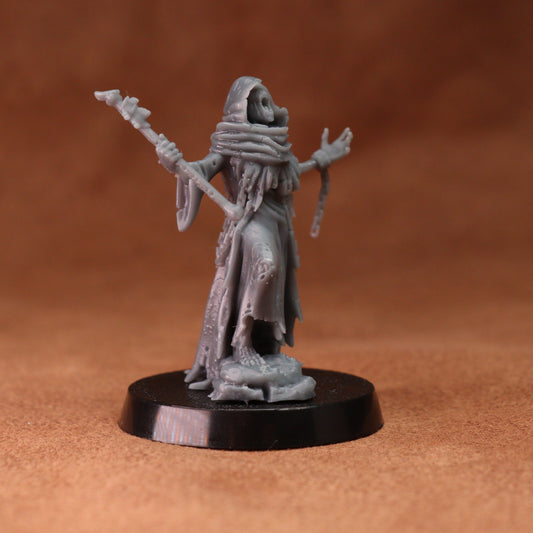 An unpainted grey tabletop gaming miniature figurine of a hooded, skeletal figure holding a staff, standing on a round black base against a warm brown textured background.