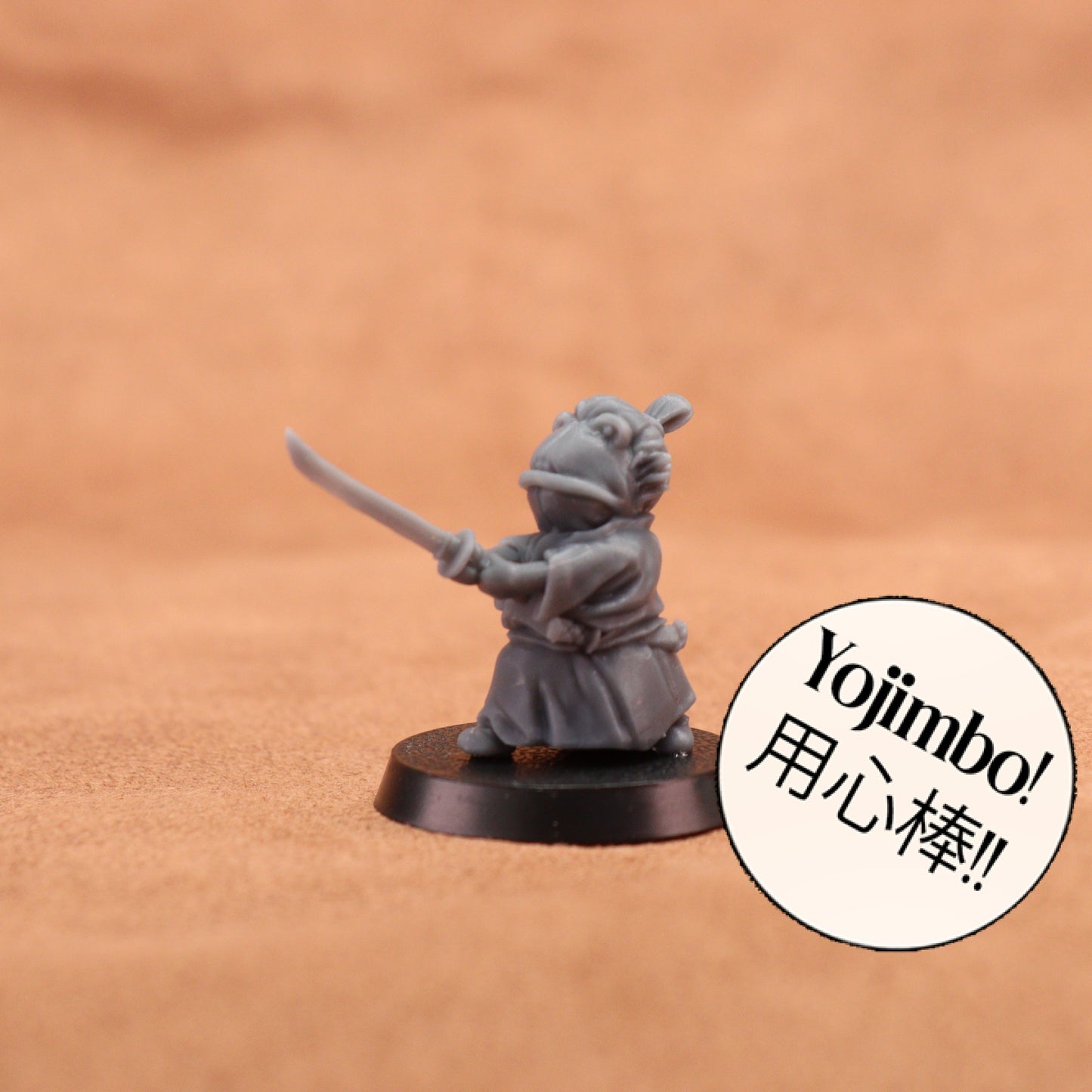 A small gray painted tabletop miniature figurine of a frog-like character in a samurai outfit holding a sword, mounted on a round black base. The background is a warm tan textured surface.