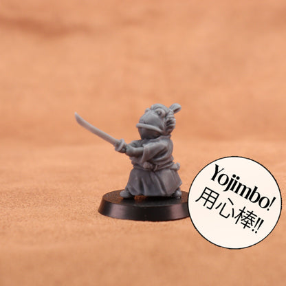 A small gray painted tabletop miniature figurine of a frog-like character in a samurai outfit holding a sword, mounted on a round black base. The background is a warm tan textured surface.