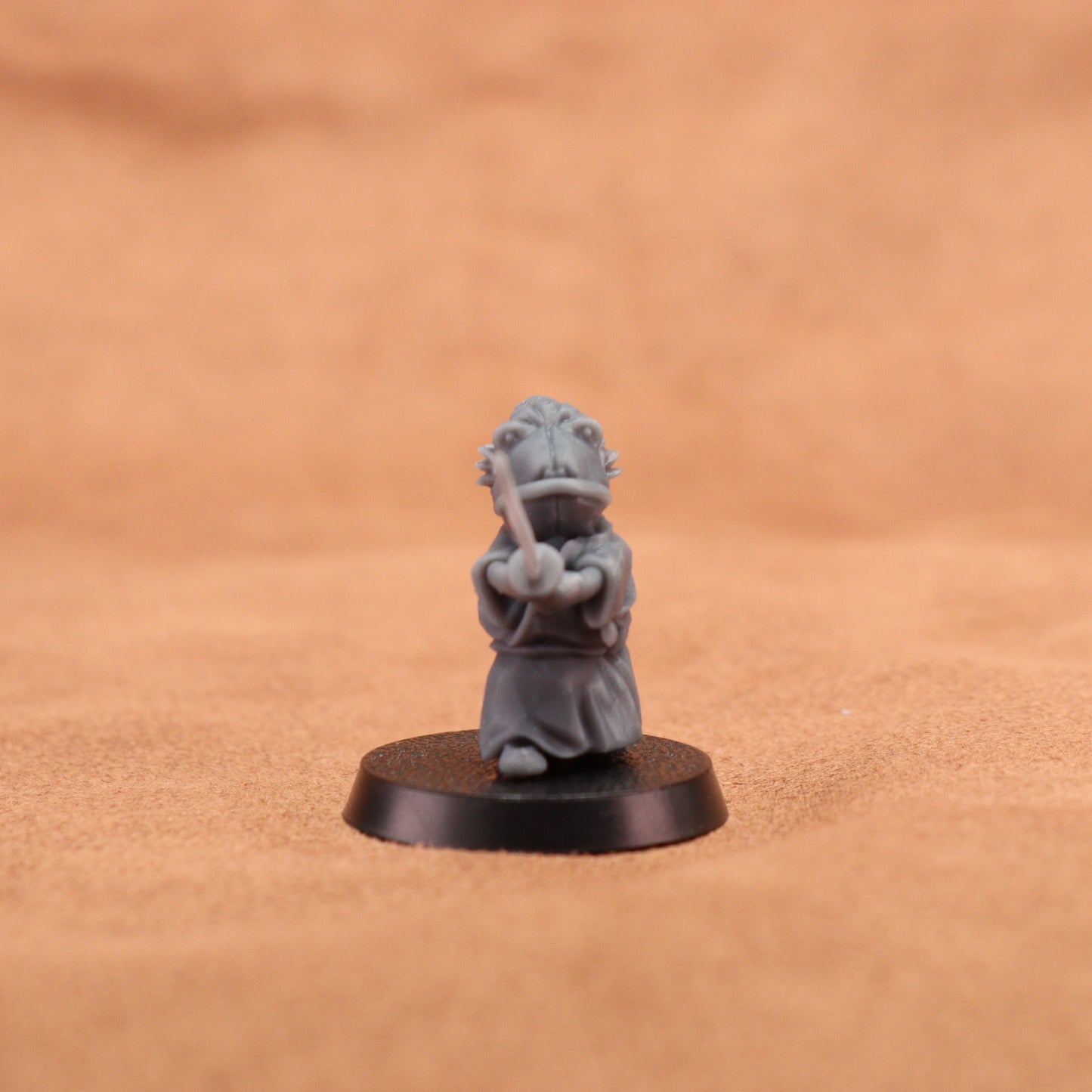 A small gray painted tabletop miniature figurine of a frog-like character in a samurai outfit holding a sword, mounted on a round black base. The background is a warm tan textured surface.
