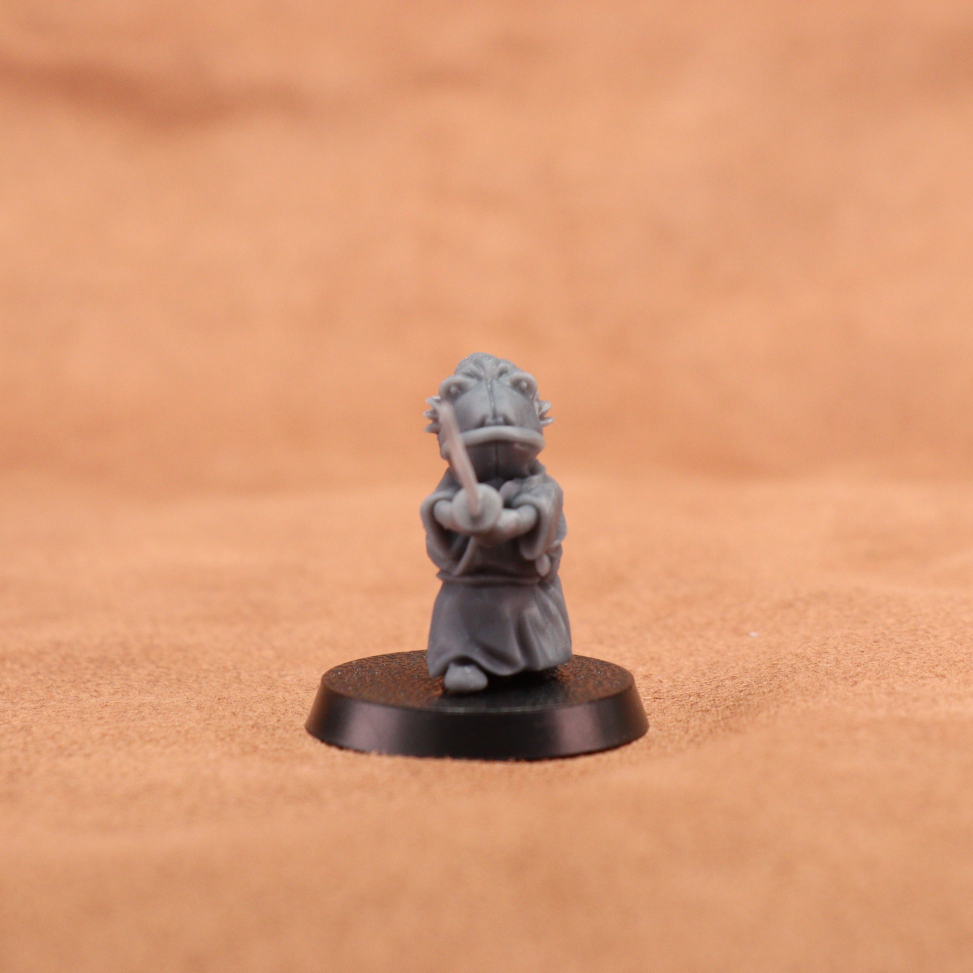 A small gray painted tabletop miniature figurine of a frog-like character in a samurai outfit holding a sword, mounted on a round black base. The background is a warm tan textured surface.