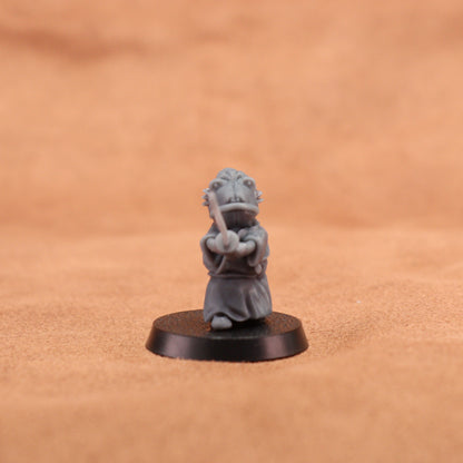 A small gray painted tabletop miniature figurine of a frog-like character in a samurai outfit holding a sword, mounted on a round black base. The background is a warm tan textured surface.