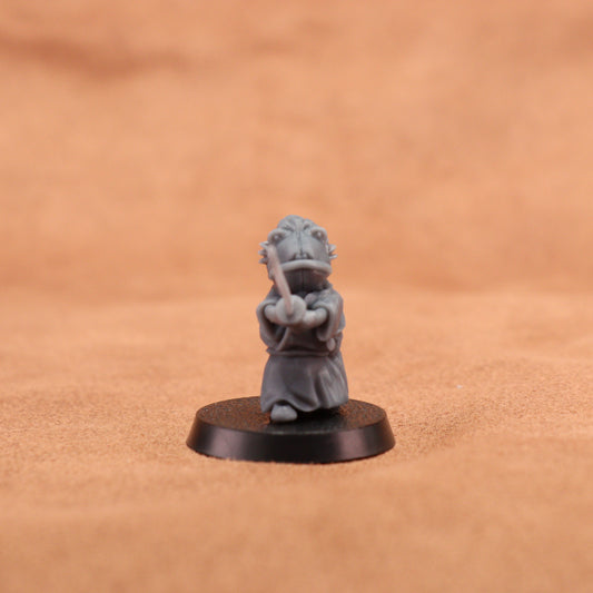 A small gray painted tabletop miniature figurine of a frog-like character in a samurai outfit holding a sword, mounted on a round black base. The background is a warm tan textured surface.