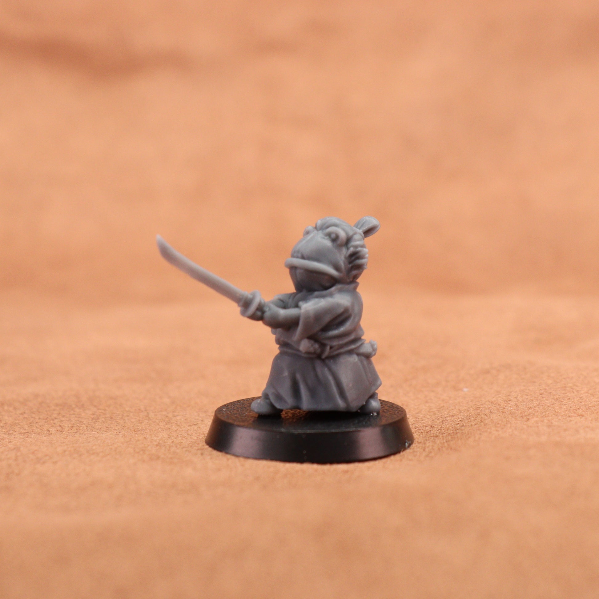 A small gray fantasy figurine of a frog-like character in a samurai-style outfit holding a sword, mounted on a round black base against a warm tan textured background.