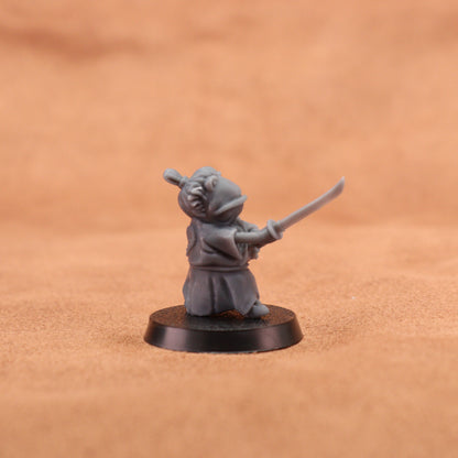 A small gray painted tabletop miniature figurine of a frog-like character in a samurai outfit holding a sword, mounted on a round black base. The background is a warm tan textured surface.