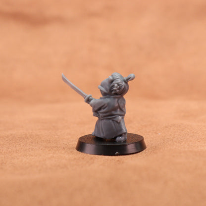 A small unpainted gray fantasy miniature figurine of a robed character holding a curved sword, mounted on a round black base against a warm tan textured background.