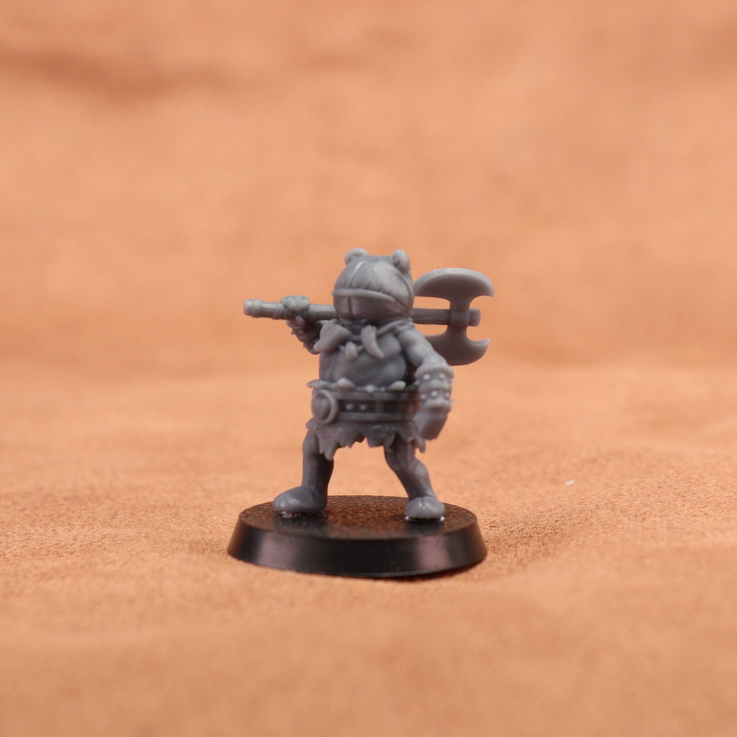 A small unpainted gray fantasy warrior miniature figurine holding a double-headed axe, standing on a round black base against a warm brown textured background.